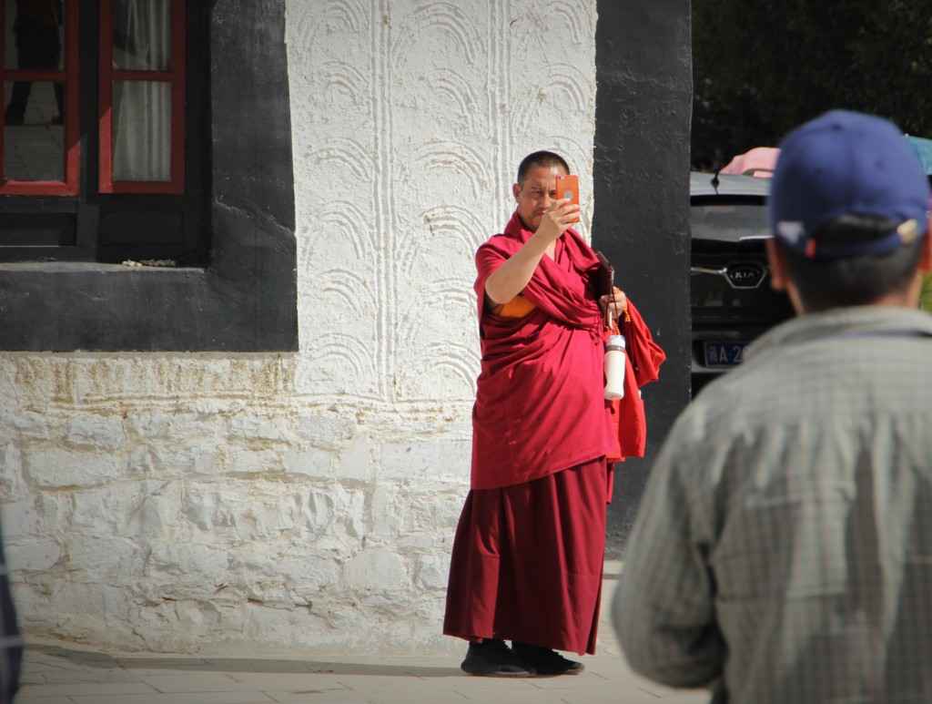 Tibetan monk taking a picture with his smartphone, while it looks like he is also carrying a tablet.